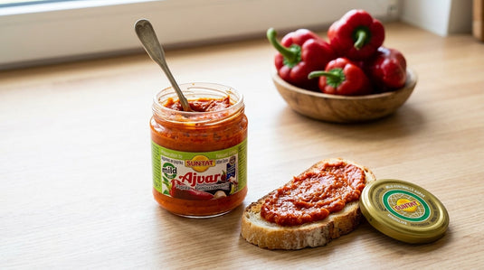 Bocal d'Ajvar doux SUNTAT ouvert avec une cuillère, tartine de pain grillé à côté et poivrons rouges en arrière-plan