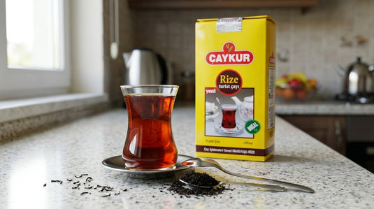 Verre à thé turc rempli de çay fraîchement infusé sur une soucoupe, avec le paquet de thé noir CAYKUR Rize et des feuilles de thé en vrac sur une cuillère, sur un plan de travail de cuisine moderne