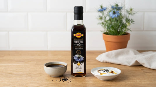 Bouteille d'huile de nigelle SUNTAT 245ml posée entre un bol d'huile et un yaourt nappé d'huile de nigelle sur un plan de travail en bois