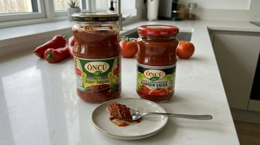 Deux pots de salça ÖNCÜ, tomate-poivron et poivron piquant pur, posés sur un plan de travail de cuisine avec une cuillère et des légumes