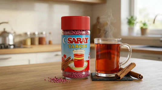 Pot de thé instantané cannelle SARAY (bouchon rouge, granulés roses) posé à côté d'une tasse de thé fumante et de bâtons de cannelle sur un plan de travail en bois
