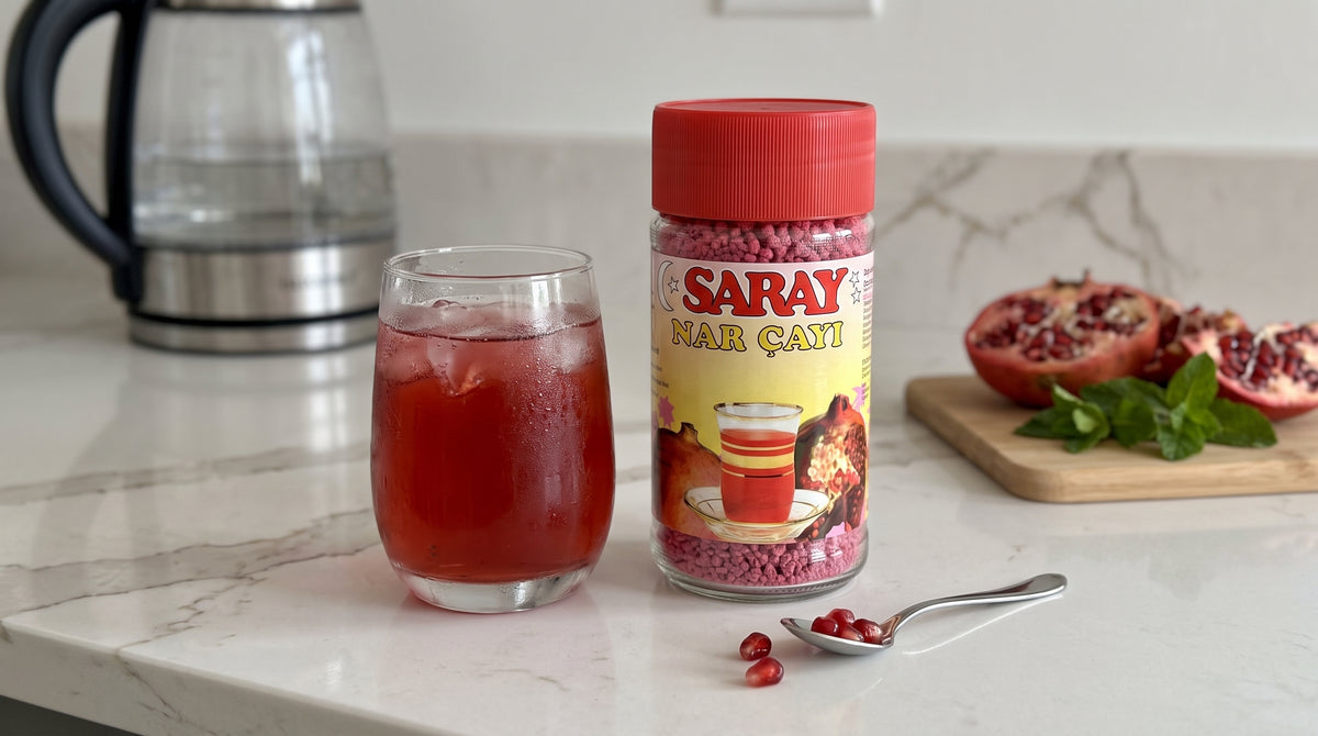Pot de thé instantané grenade SARAY posé à côté d'un verre de thé glacé rosé, d'une grenade coupée et de menthe fraîche, sur un plan de travail de cuisine moderne